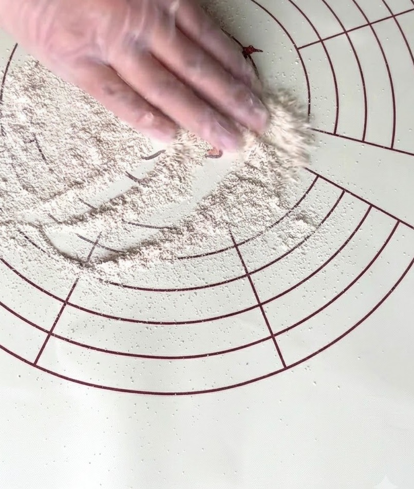 Una mano guantata che sparge farina integrale su un tappetino da cucina circolare in silicone.