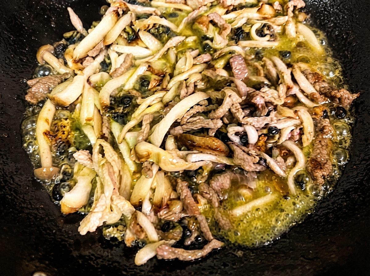 Funghi shiitake tagliati a julienne, funghi enoki e lumache di mare che soffriggono nellolio caldo allinterno di un wok nero.
