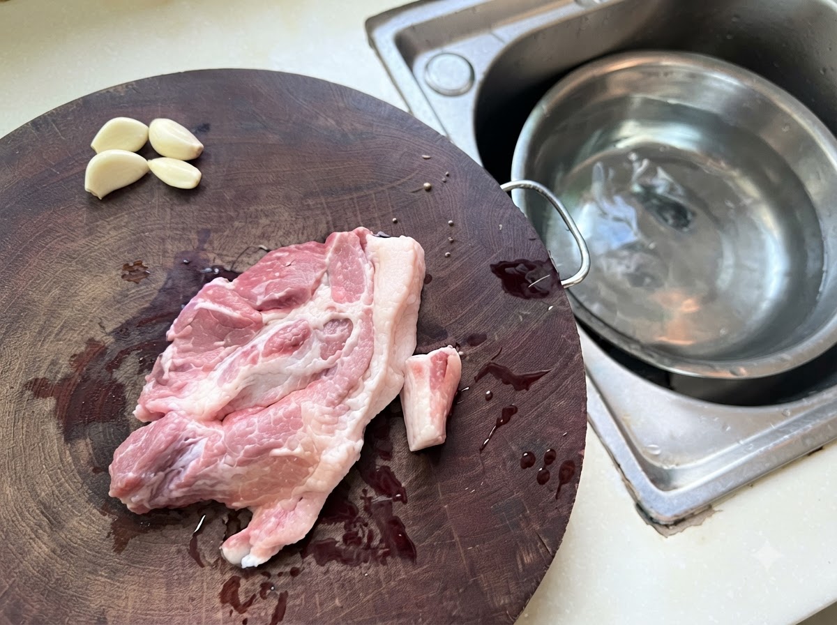Pezzi di maiale macinato e aglio sparsi su un tagliere di legno accanto a una ciotola di metallo con acqua.
