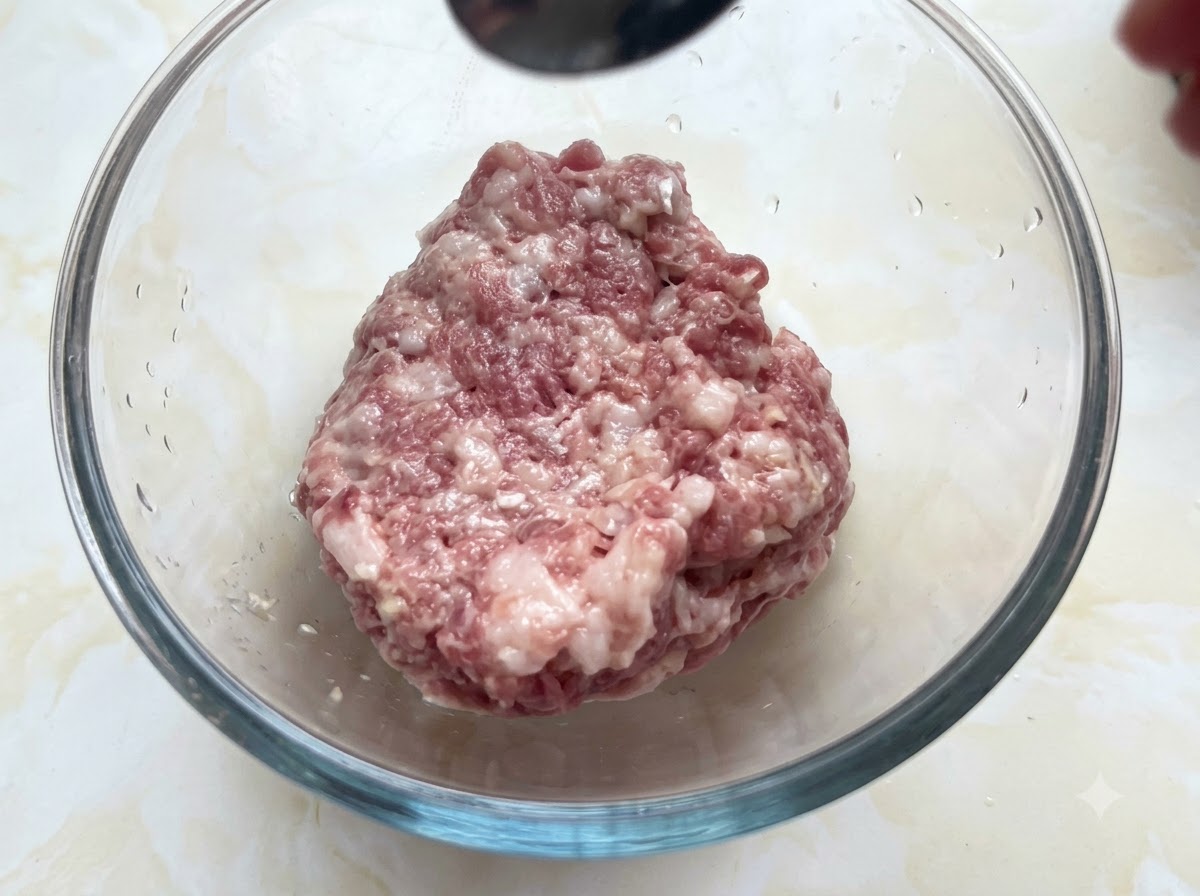 Una ciotola di vetro riempita di maiale macinato che riceve i condimenti da un cucchiaio tenuto sopra.
