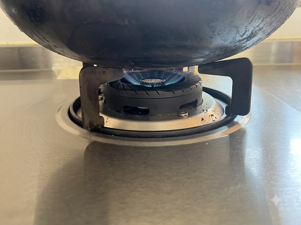 Una fiamma blu bassa che riscalda il fondo di un wok nero su un piano cottura in acciaio inossidabile.