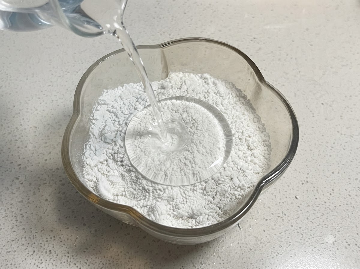 Acqua versata da un misurino in una ciotola di vetro contenente un mix di farina e amido.