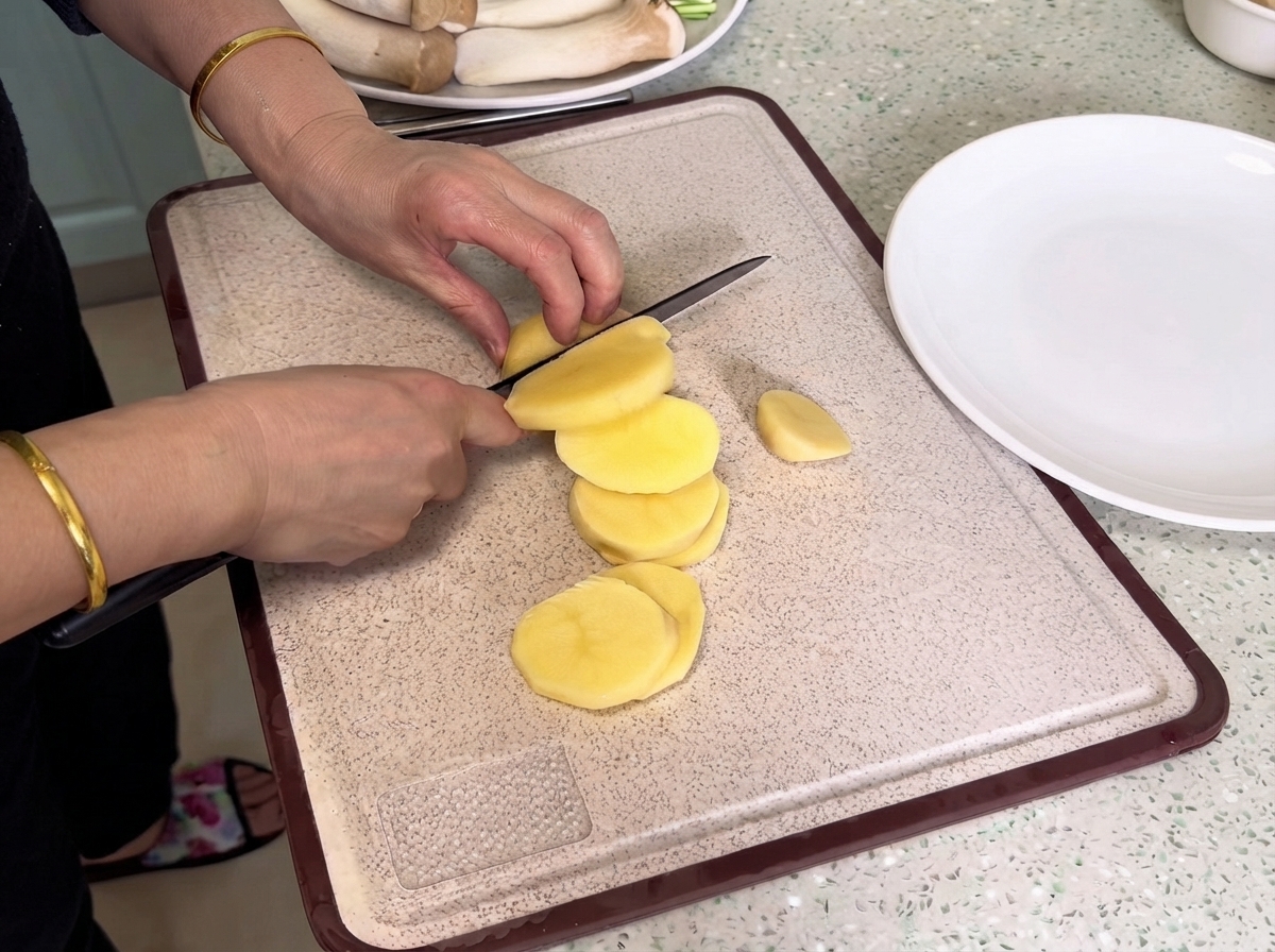 Patate affettate con un coltello su un tagliere vicino a un piatto bianco.