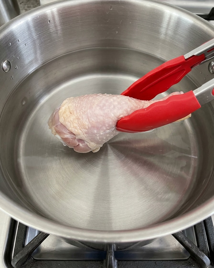 Una coscia di pollo viene inserita con le pinze in una pentola di acciaio inossidabile piena di acqua fredda.