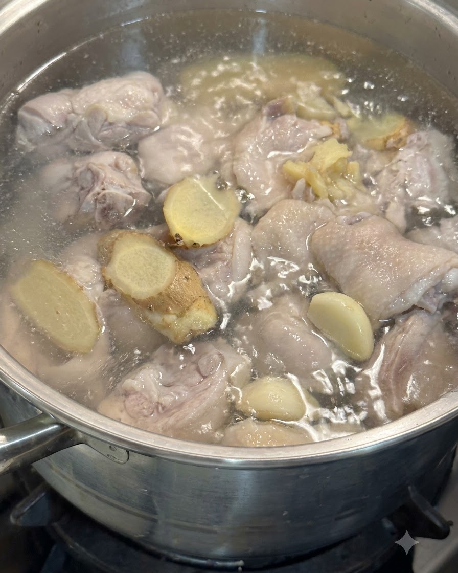 Spicchi daglio e fette di zenzero che galleggiano nella pentola con i pezzi di pollo in acqua sobbollente.