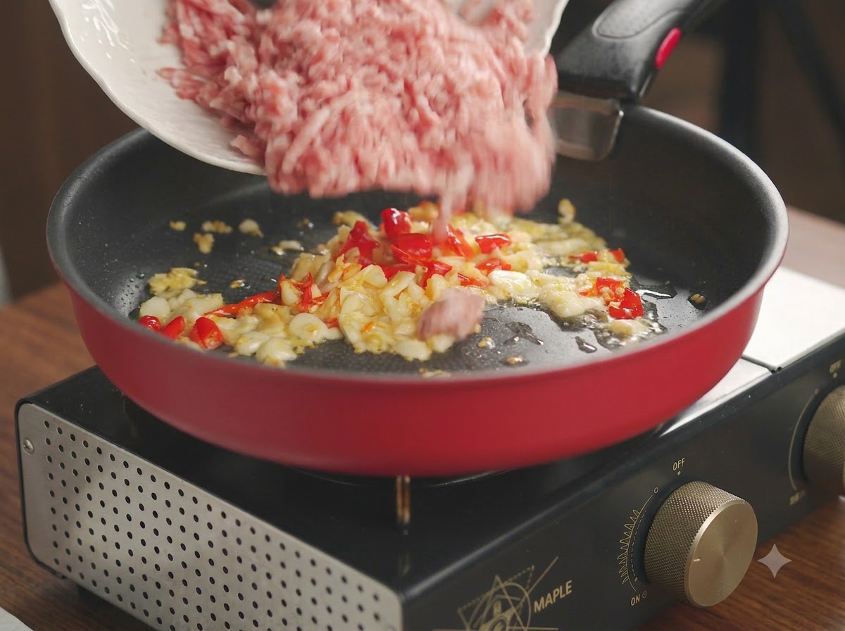 Carne di maiale macinata cruda versata da un piatto bianco in una padella rossa con aglio e peperoncino soffritti.