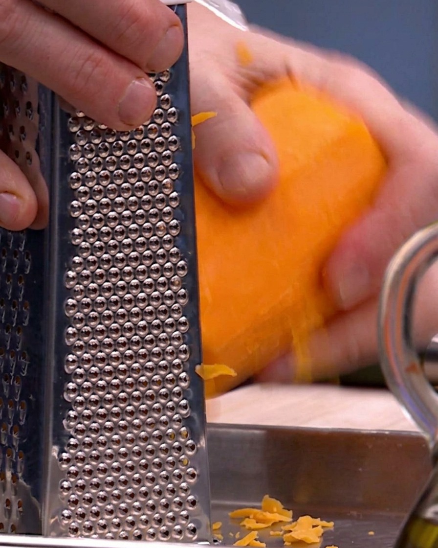 Mani che usano una grattugia a scatola di metallo per sminuzzare la zucca butternut arancione brillante su un vassoio di metallo.