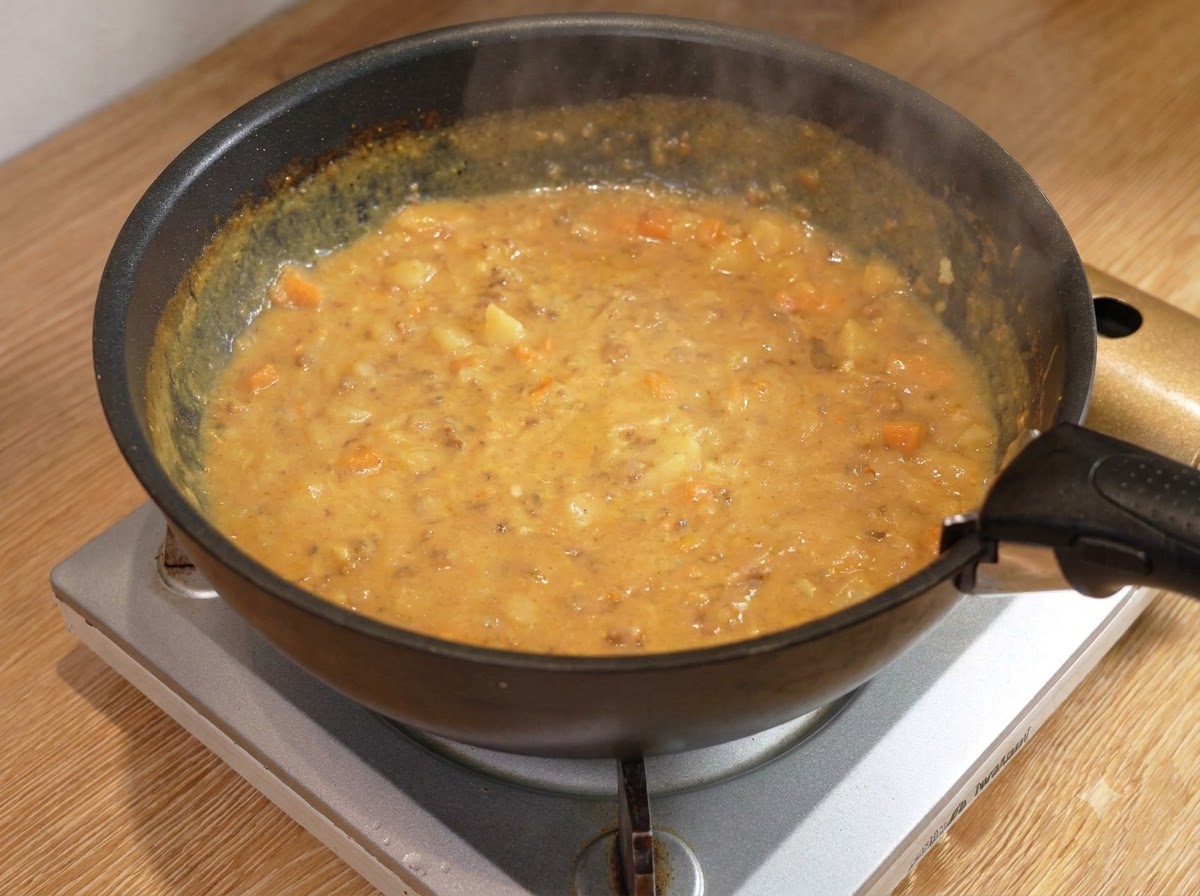 Un cucchiaio di legno che mescola una densa e bollente salsa di carne al curry allinterno di una padella nera su un fornello.