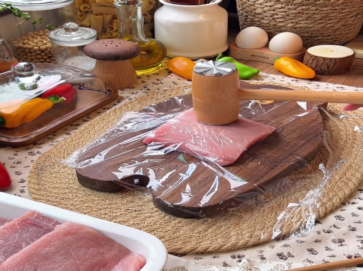 Un batticarne di legno che batte su un pezzo di filetto di maiale coperto di pellicola trasparente su un tagliere di legno.