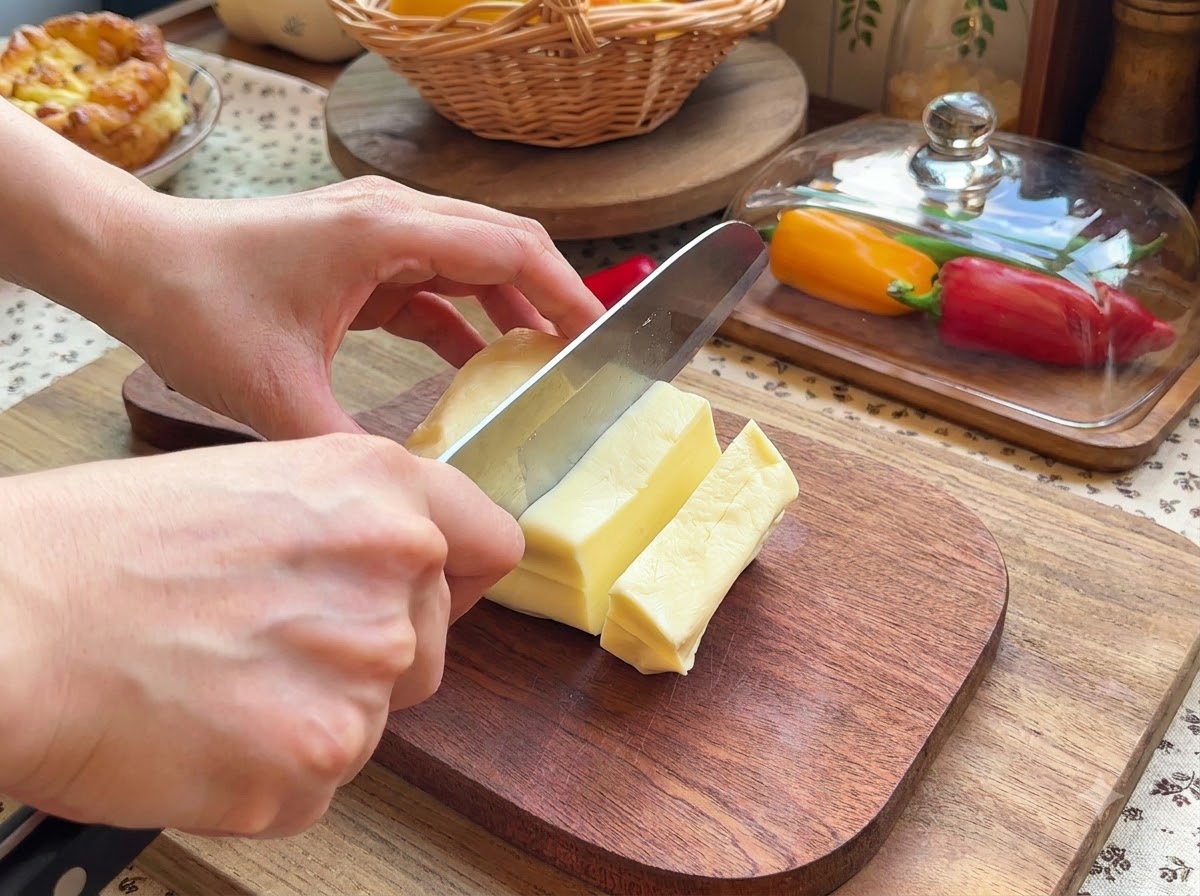 Un paio di mani che usano un coltello per affettare un blocco di mozzarella in piccoli pezzi rettangolari su un tagliere di legno.