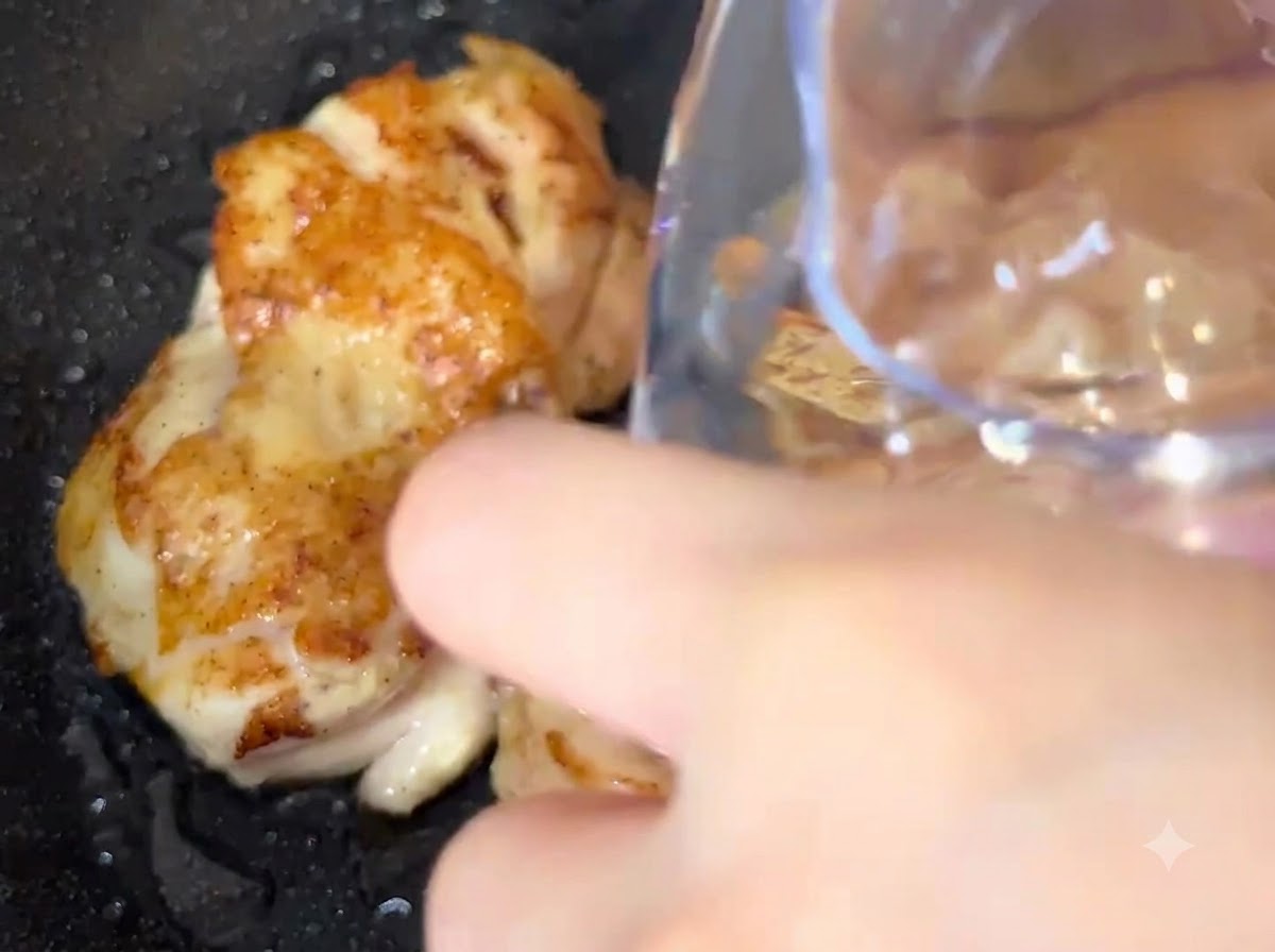 Acqua versata da un contenitore direttamente in una padella con pezzi di pollo in cottura.