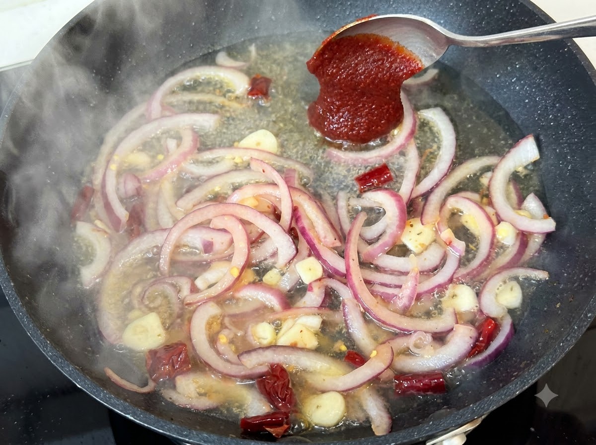Un cucchiaio di pasta di peperoncino coreano aggiunto al liquido della padella in ebollizione con cipolle e spezie.