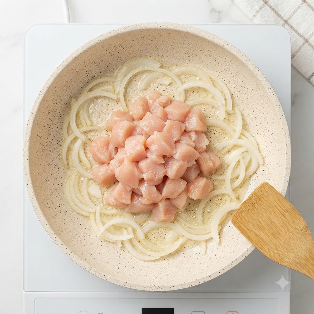 Petto di pollo crudo a cubetti ammucchiato al centro di una padella circondato da fette di cipolla saltate.