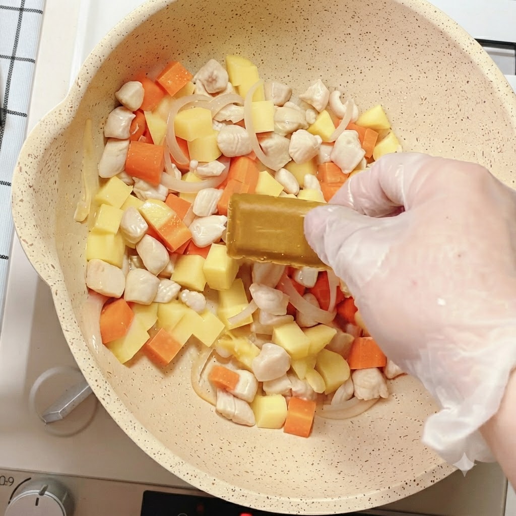 Una mano che posiziona un blocchetto di condimento al curry nella padella piena di verdure miste e pollo.