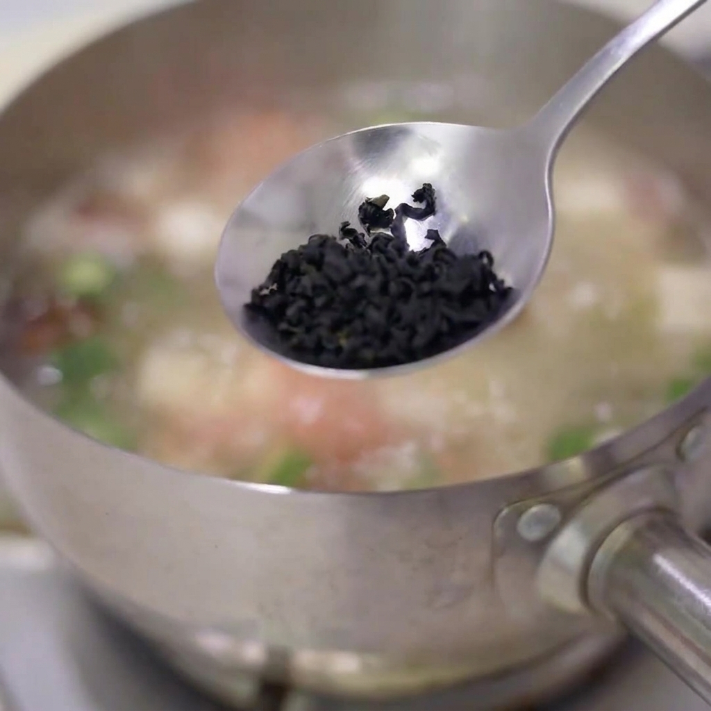 Un cucchiaio di fiocchi di alga disidratata fatto cadere in una pentola di zuppa in ebollizione in acciaio inossidabile.