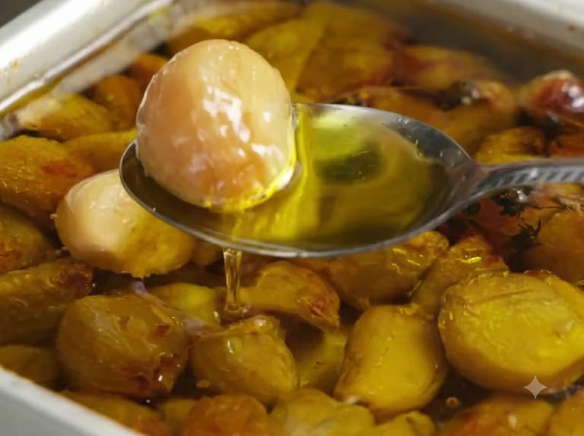 Un cucchiaio che raccoglie uno spicchio daglio arrostito dorato da una teglia piena di olio doliva e spicchi daglio.