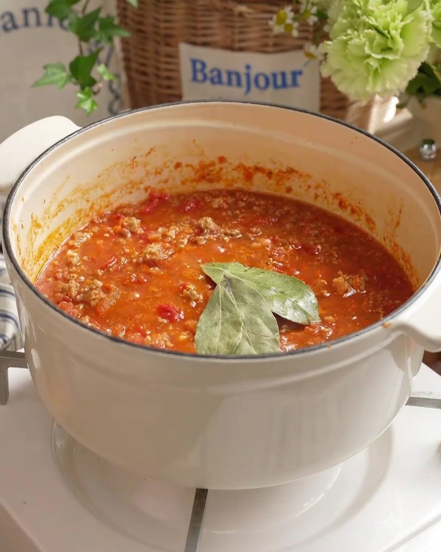 Foglie di alloro e acqua fresca aggiunte al sugo di carne di pomodoro che sobbolle in una pentola bianca.