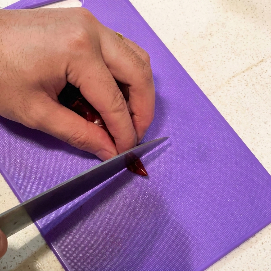Affettare spicchi daglio e tagliare peperoncini secchi a pezzi su un tagliere viola.