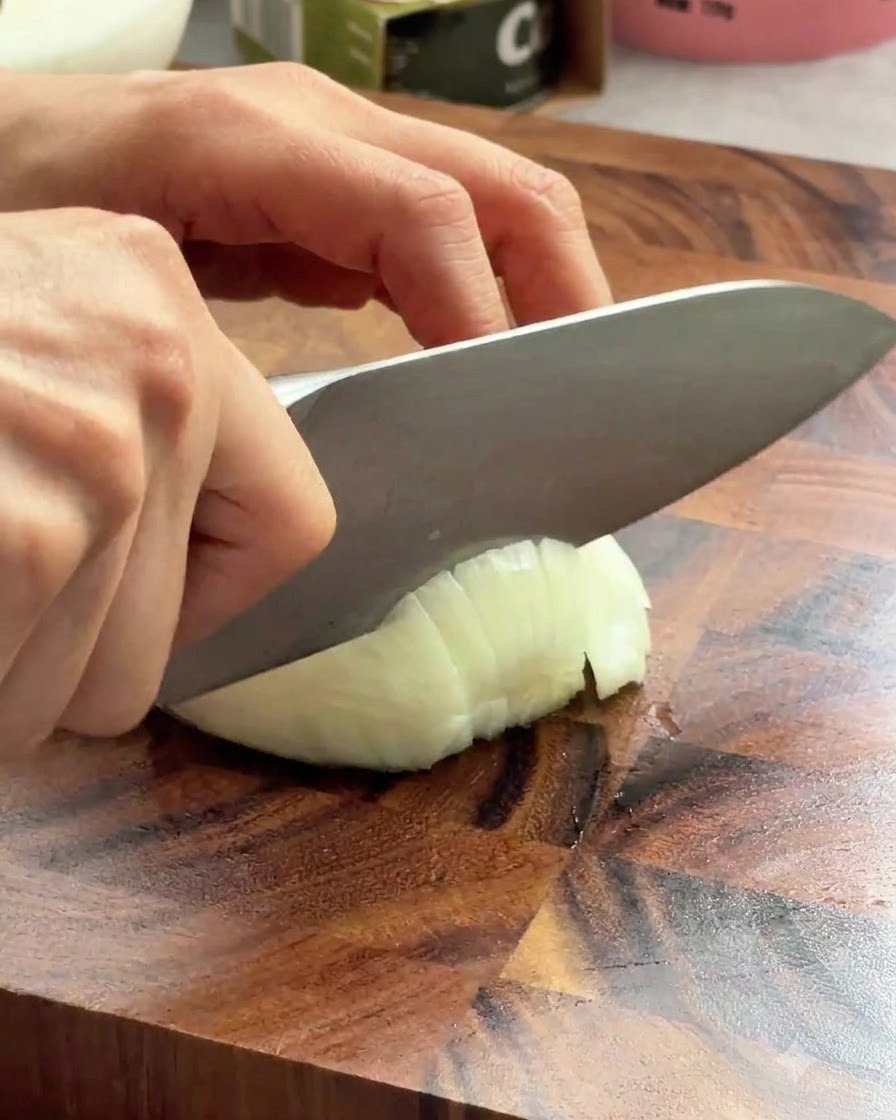 Un coltello da chef affilato che affetta una cipolla bianca su un asse di legno.