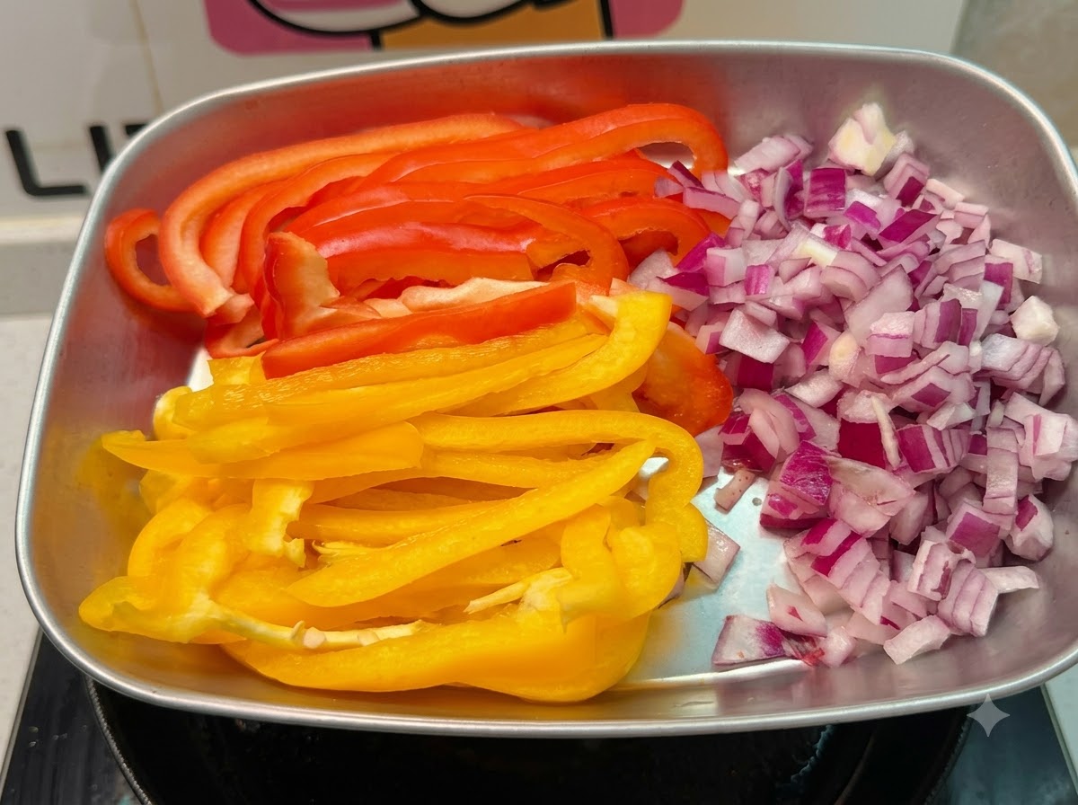 Una teglia di metallo con cipolle rosse tagliate a dadini e strisce lunghe di peperoni rossi e gialli.