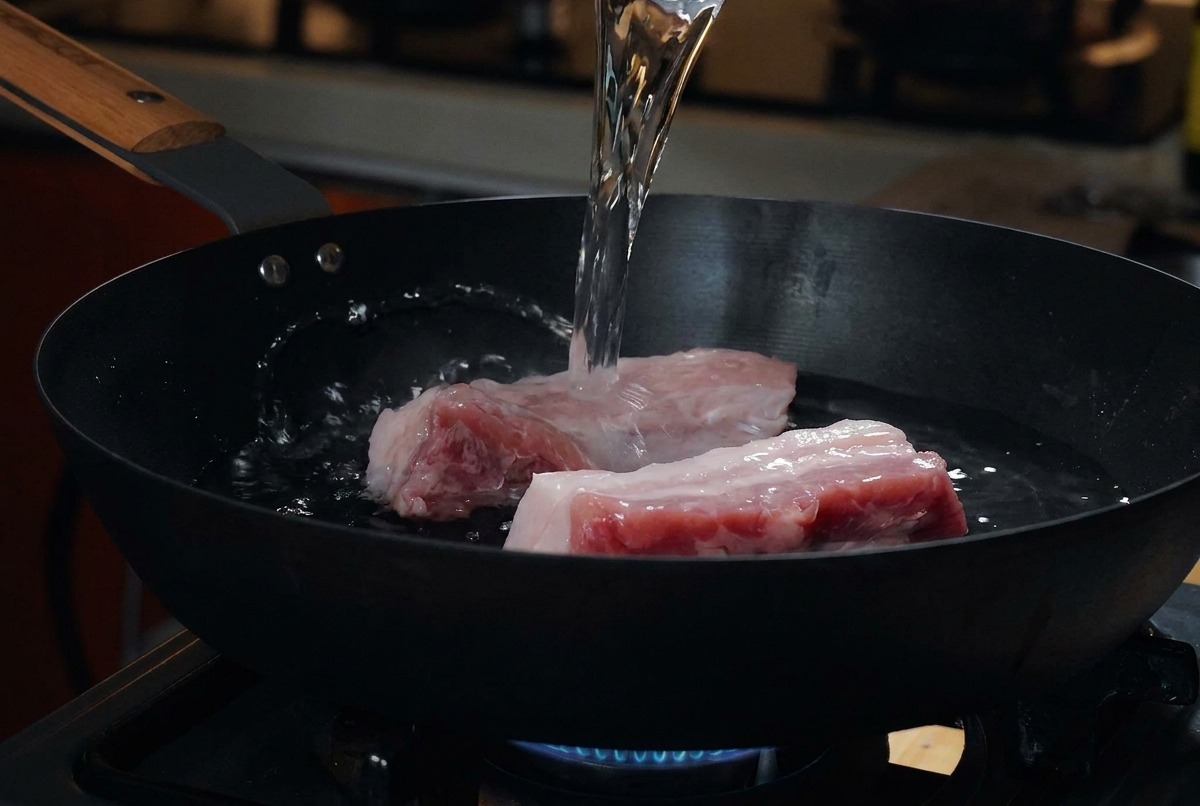 Spessi pezzi di pancetta di maiale cruda riposano in una padella nera fredda mentre viene versata dellacqua da un misurino.