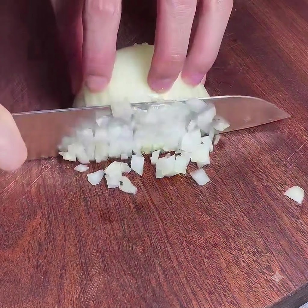 Mani che tengono un coltello per tritare finemente una cipolla su un tagliere di legno.