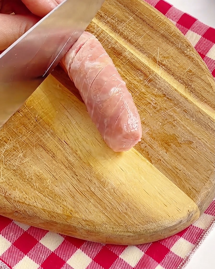 Una mano che incide con cura la superficie di una salsiccia rosa intera con un coltello su un tagliere di legno.