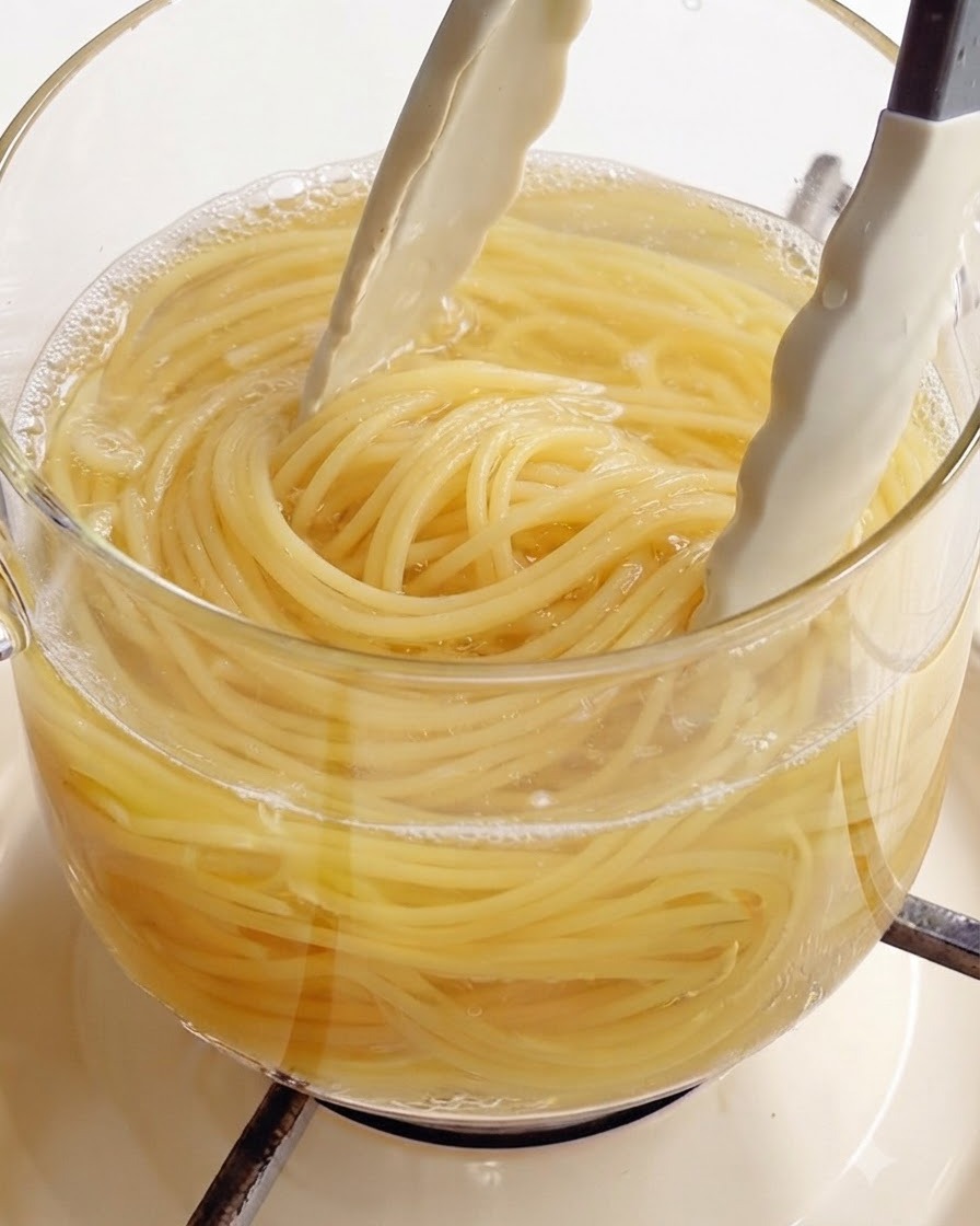Utilizzo di pinze da cucina bianche per sollevare spaghetti perfettamente cotti da una pentola di vetro trasparente di acqua bollente.