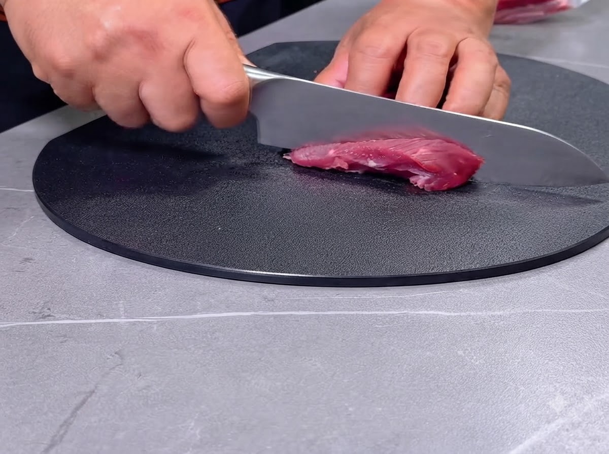 Una persona che taglia carne rossa cruda a pezzi con un coltello su un tagliere nero.
