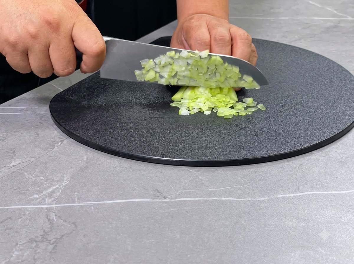 Un paio di mani che usano un coltello da chef per tagliare a dadini gambi di sedano verde su un tagliere nero circolare.