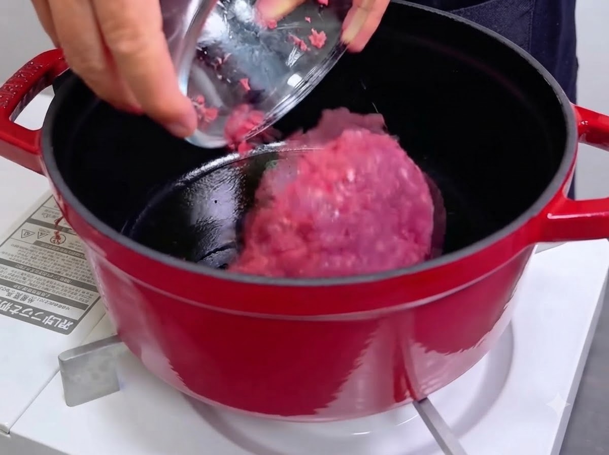 Una ciotola che versa carne macinata cruda e rosa in una pentola di ghisa rossa calda.