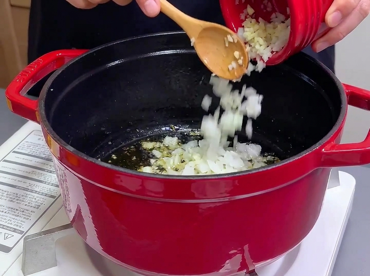 Versare cipolle e aglio tritati da una ciotola rossa piccola in una pentola di ghisa con olio caldo.