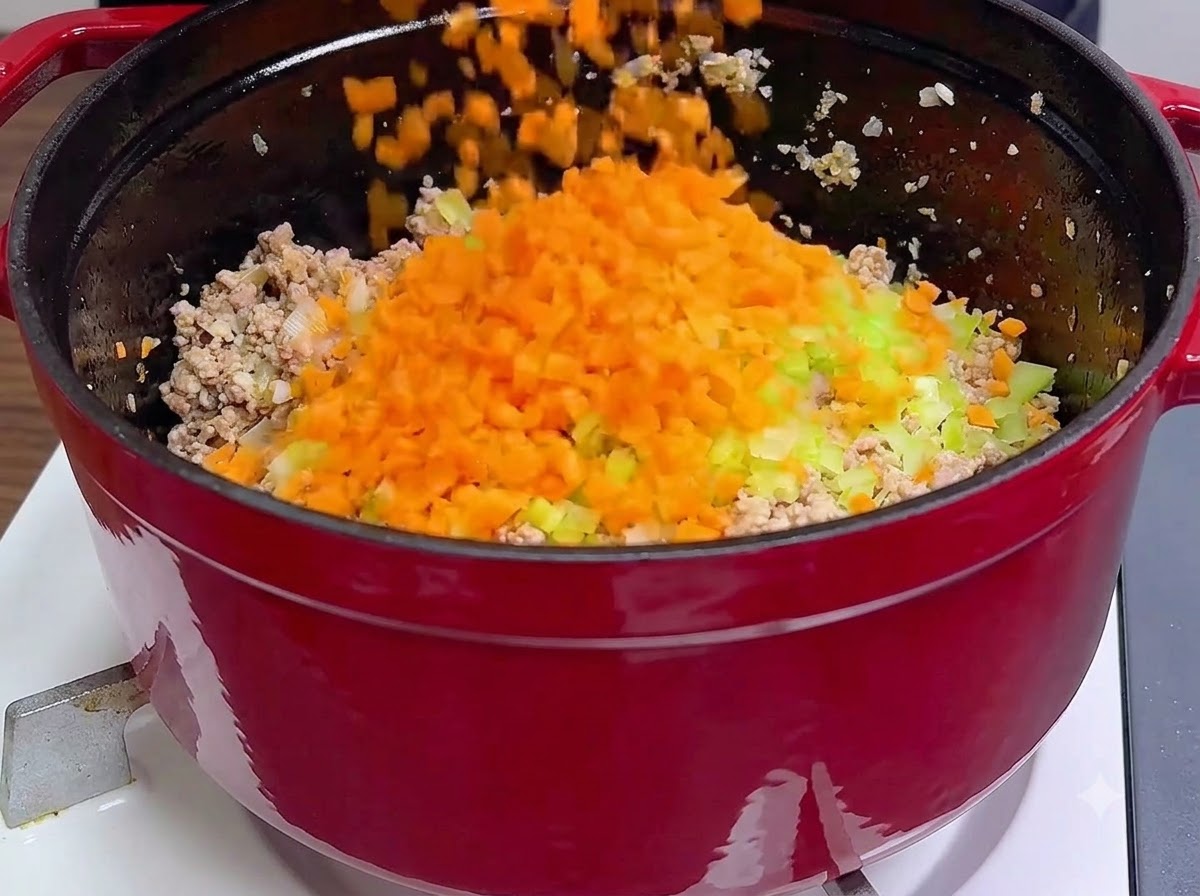 Carote arancioni e sedano verde tritati freschi versati direttamente sopra la carne macinata cotta allinterno di una pentola rossa.