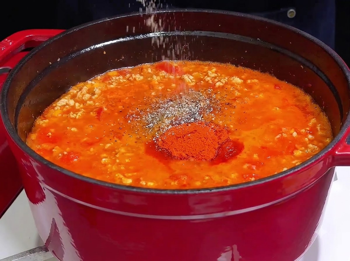 Una mano che cosparge sale e spezie sopra un sugo di carne rosso vibrante e sobbollente in una pentola di ghisa.