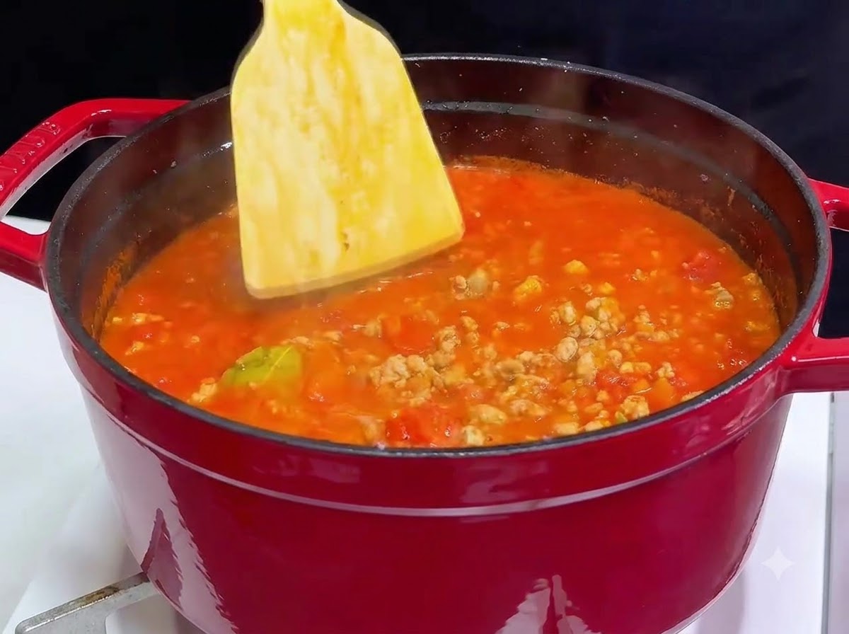 Una spatola di legno che mescola il vino rosso in una pentola di ghisa rossa piena di salsa di carne e pomodoro bollente.