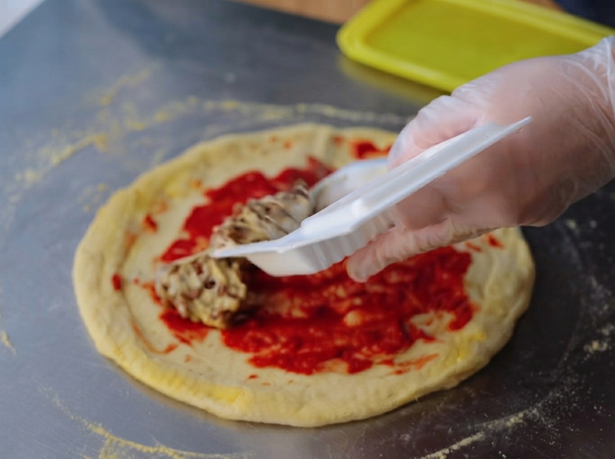 Una mano guantata che spalma un composto di natto appiccicoso e filamentoso in modo uniforme sopra lo strato di concentrato di pomodoro rosso vivo su una base di pizza cruda.