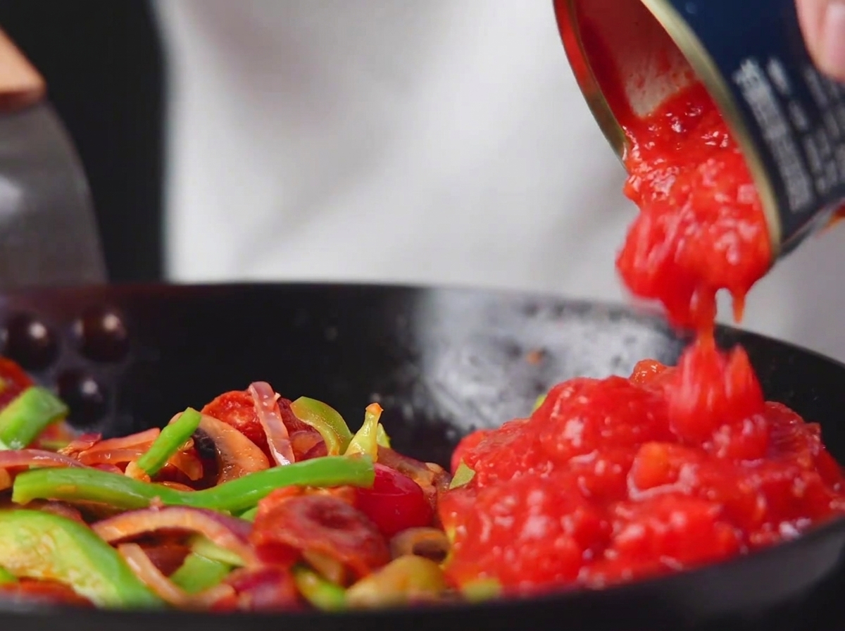 Polpa di pomodoro versata nella padella con verdure e salame.