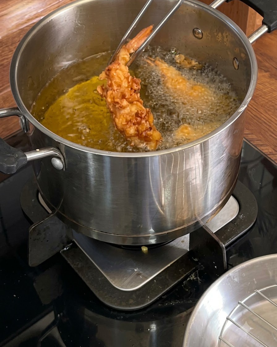 Pinze di metallo che tengono un gambero fritto croccante e dorato sopra una pentola calda di olio.