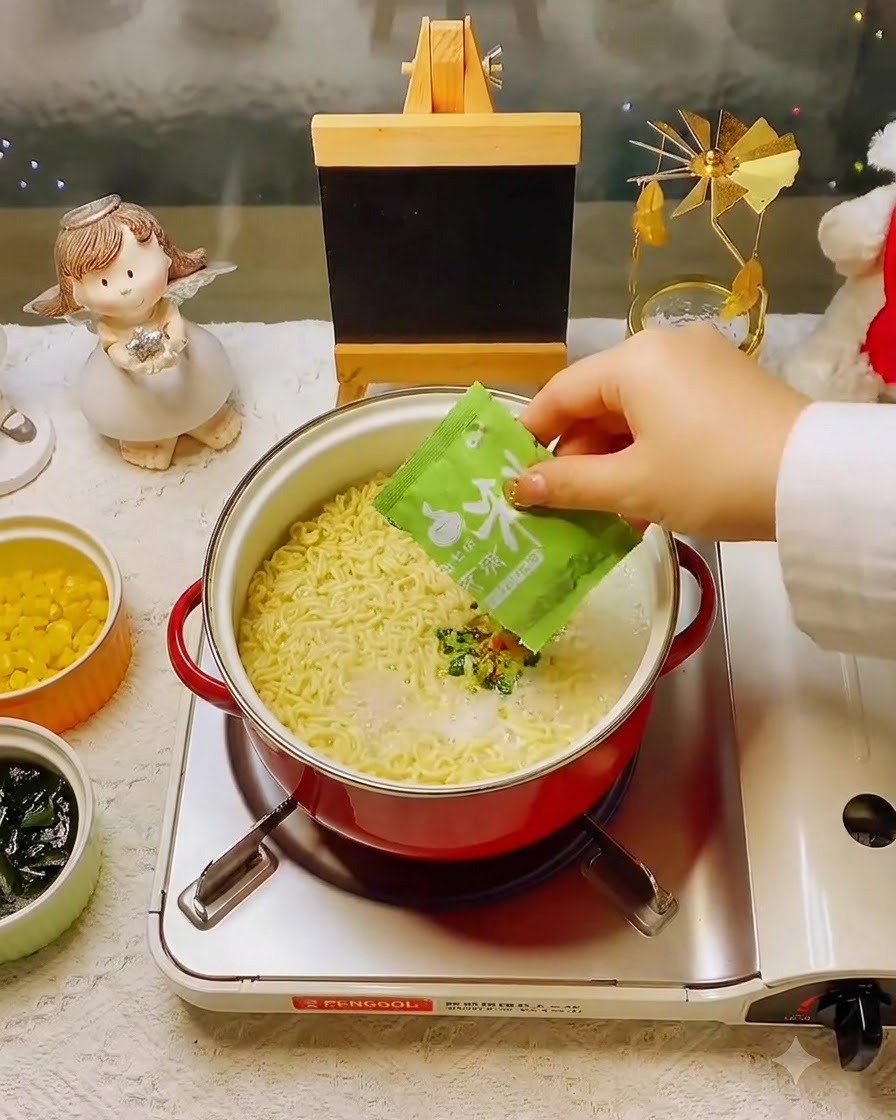 Una mano che aggiunge una bustina di condimento verde nella pentola di noodles e brodo.