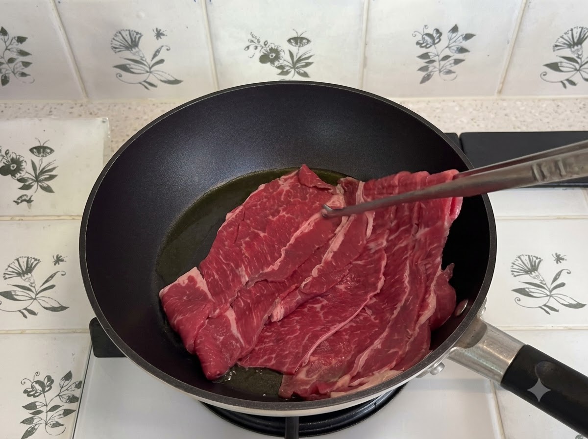 Manzo grasso affettato sottilmente fatto saltare in una padella scura con pinze da cucina.