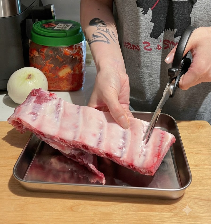 Mani che usano forbici da cucina per tagliare un grosso rack di costine di maiale crude in pezzi singoli su un tagliere di legno.