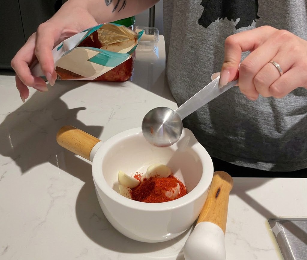 Misurazione del peperoncino rosso in polvere in un mortaio bianco riempito di spicchi daglio.