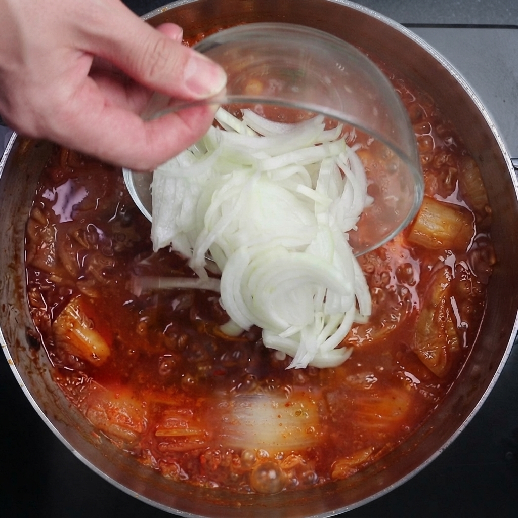 Cipolle bianche affettate versate da una ciotola di vetro in una pentola di brodo di kimchi rosso e piccante che bolle vigorosamente.