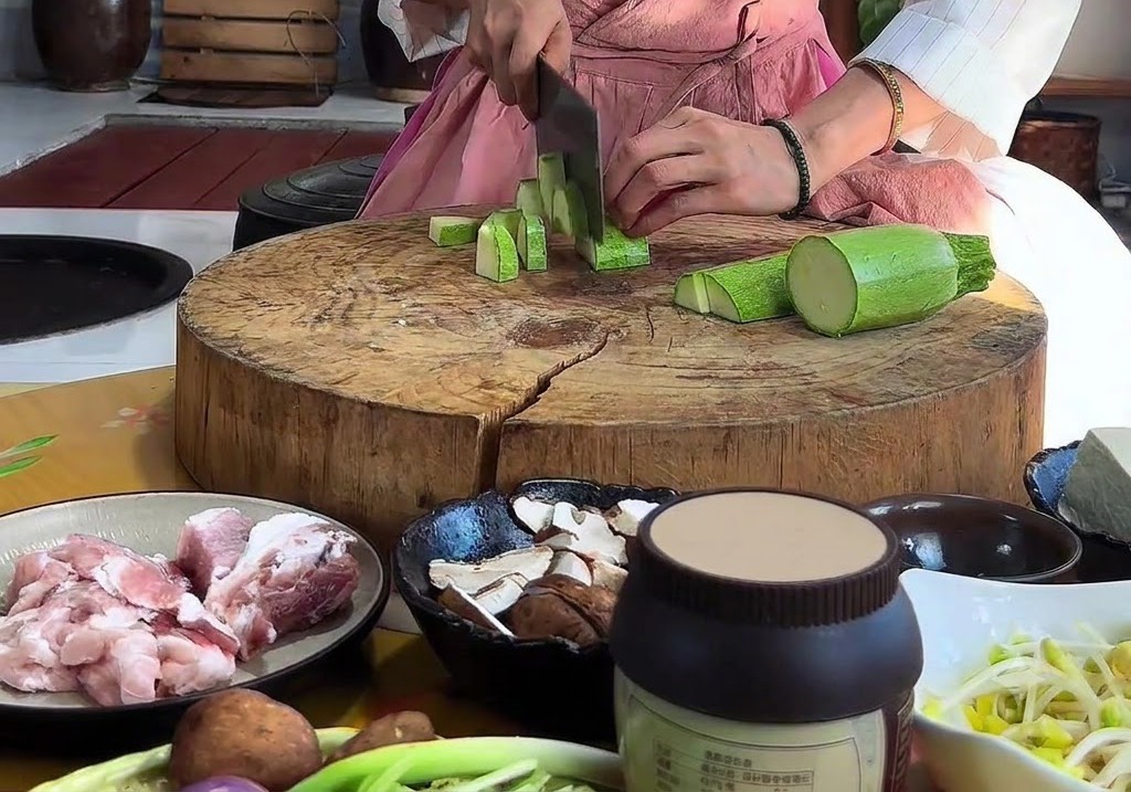 Mani che tagliano una zucchina verde a triangoli con una mannaia su un tagliere rotondo di legno.