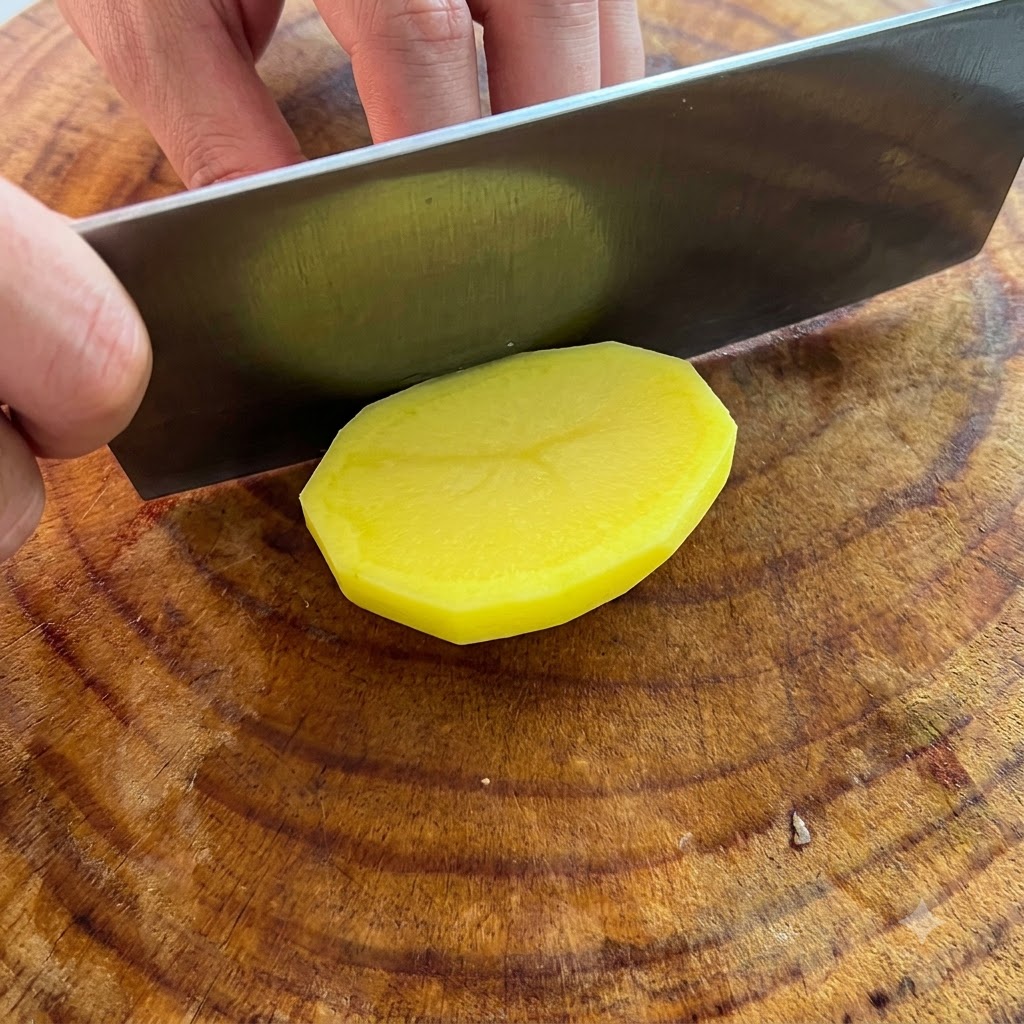 Un coltello che taglia una patata gialla sbucciata a fette spesse su un tagliere di legno.