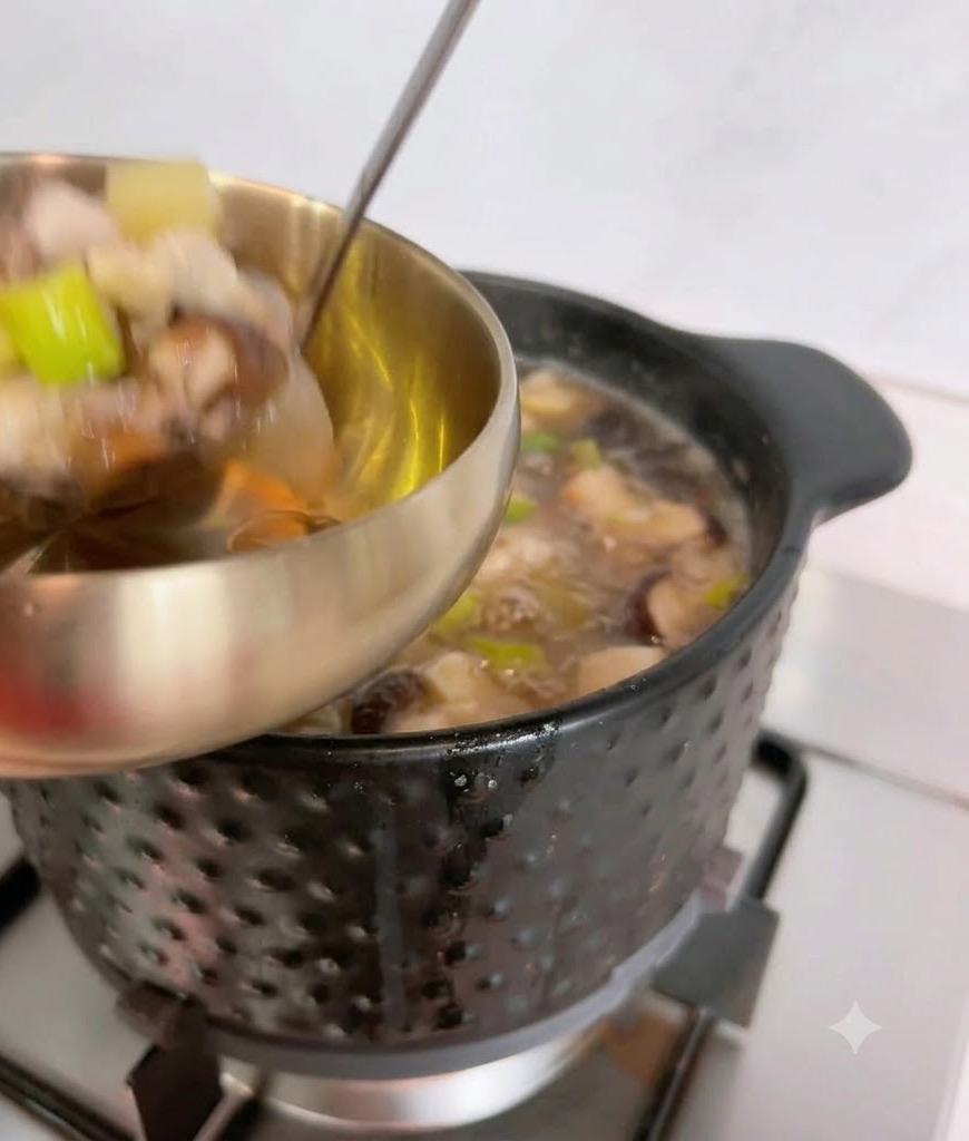 Un mestolo che raccoglie la zuppa calda di pasta di soia e i suoi ingredienti da una pentola nera in una ciotola di metallo.