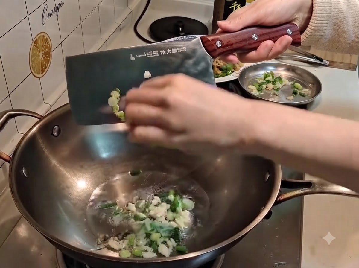 Una mano usa una mannaia per spingere cipollotti e aglio tritati nellolio caldo di un wok.
