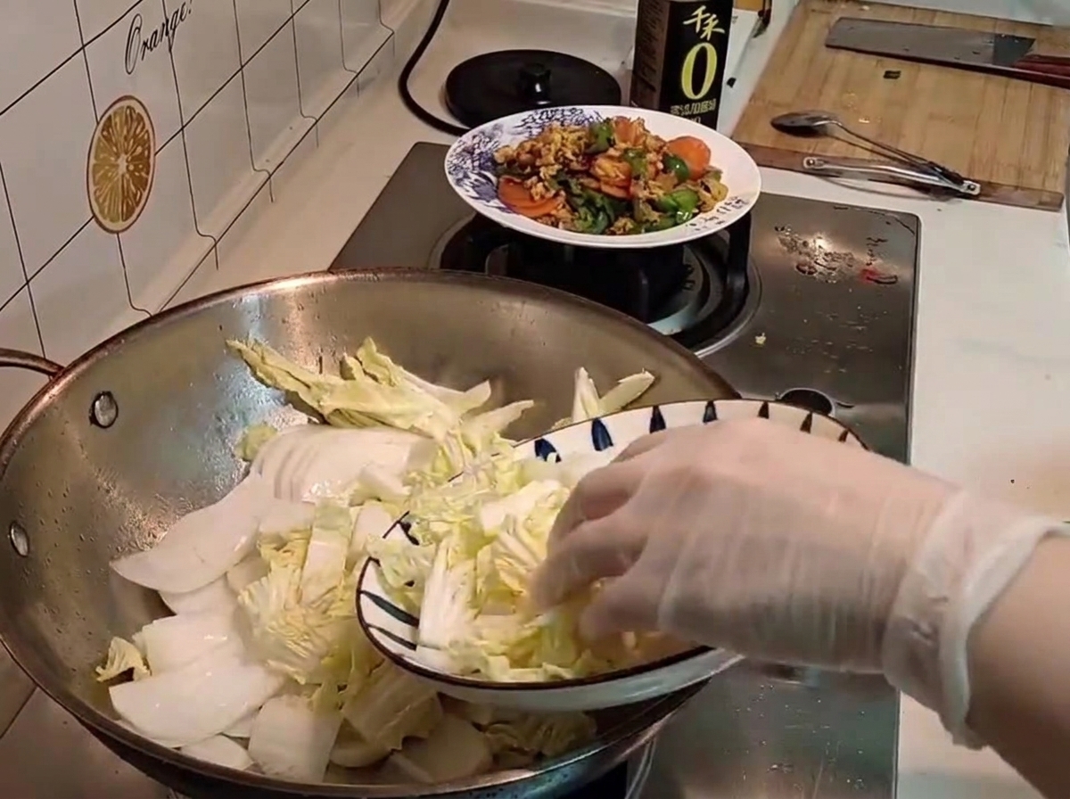 Una mano con guanto trasferisce del cavolo napa crudo affettato da un piatto decorato a un wok.