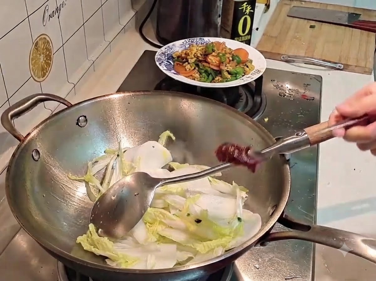 Una mano tiene un cucchiaio di metallo con pasta di peperoncino coreana rosso scuro sopra un wok pieno di cavolo e ravanello.