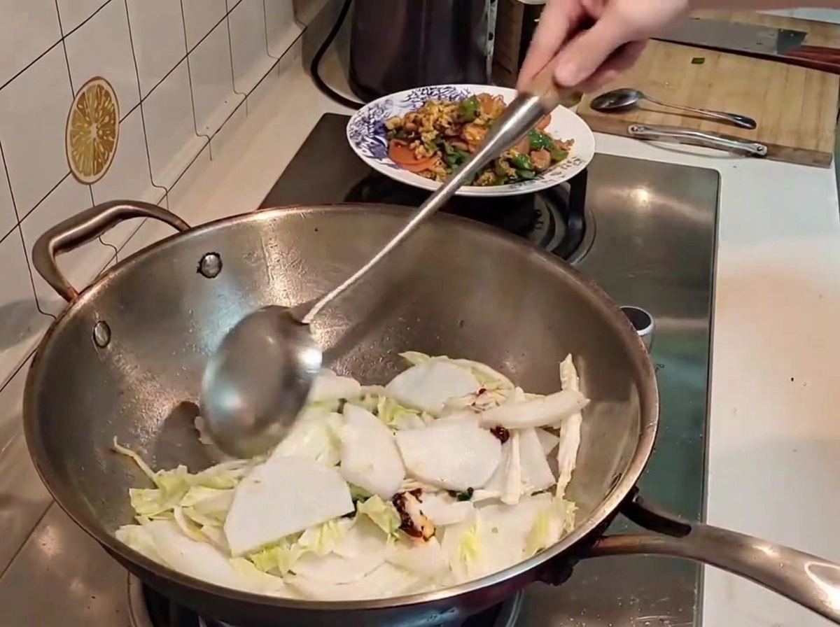 Una spatola di metallo mescola pezzi di cavolo napa e daikon con pasta di peperoncino allinterno di un wok caldo.
